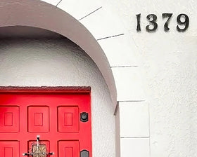 Número de casa fabricado en acero con tipografía original serif personalizada, instalado sobre muro fachada blanca con puerta roja, señalética moderna dirección domicilio fabricado a medida por IronPig Chile