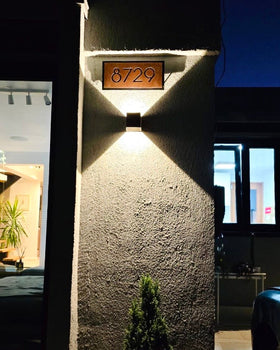 Placa de número de casa, letrero dirección en acero tipo corten oxidado con tipografía moderna, iluminada con aplique de luz cálida bidireccional en fachada exterior de noche, testimonio cliente chileno, fabricada a medida por IronPig Chile