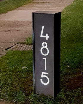 Tótem de acero negro con número de casa 4815 en tipografía moderna retroiluminada, instalado verticalmente en jardín residencial exterior, fabricado a medida por IronPig Chile