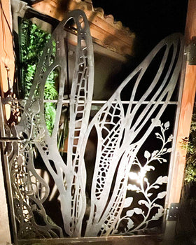 Portón de acero calado con diseño botánico de hojas y ramas entrelazadas, iluminado con luz nocturna, instalado en acceso de jardín residencial, fabricado a medida en Chile por IronPig