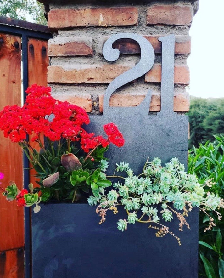 Número de casa en acero decorativol, instalado sobre muro de ladrillos, diseño de número domicilio con macetero ideal para plantas, suculentas y flores rojas, casa entrada alegre natural acogedora, fabricado por IronPig Chile. Testimonio cliente